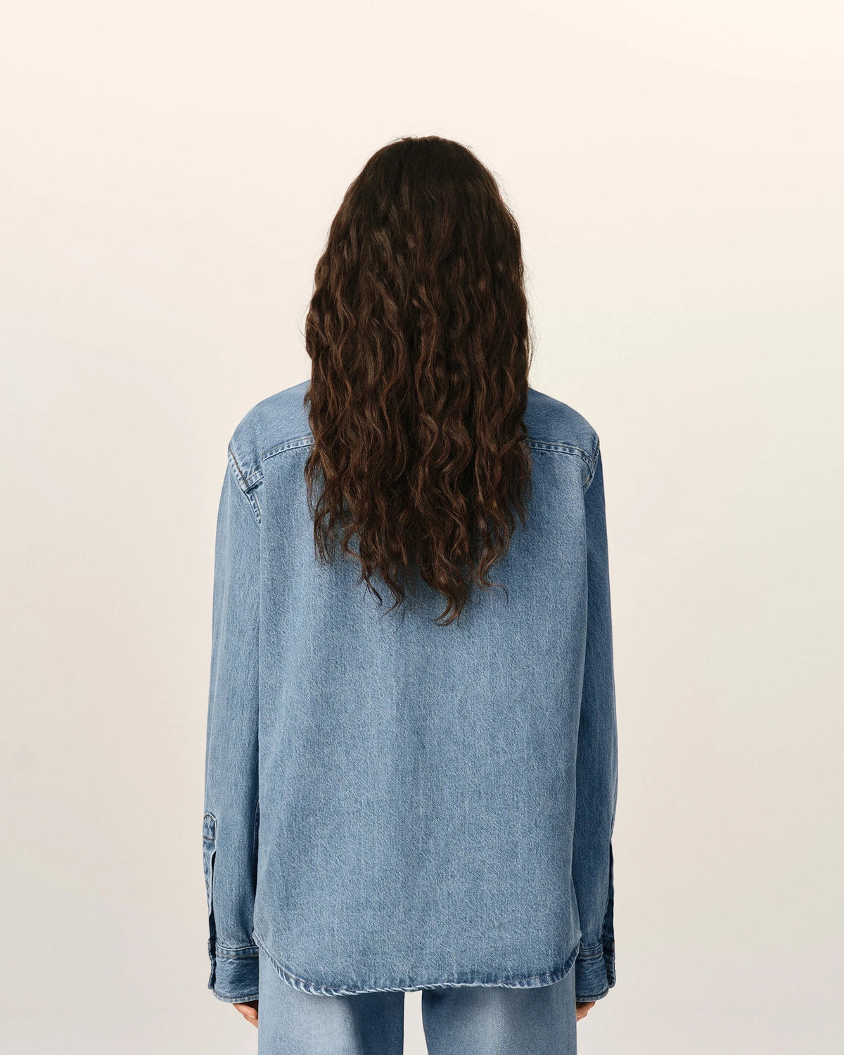 Back view of a woman with long wavy hair wearing an Ami Paris light blue denim shirt with tobacco stitching.